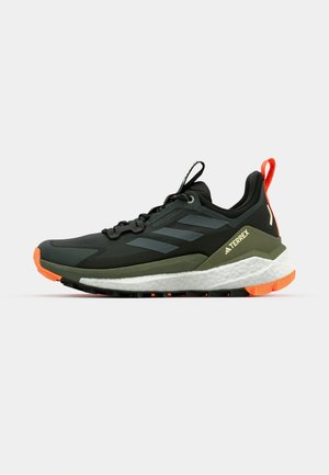 Zapatilla deportiva en negro y gris con acentos verdes, entresuela blanca y suela naranja. Presenta un diseño de cordones y una banda de rodadura texturizada.