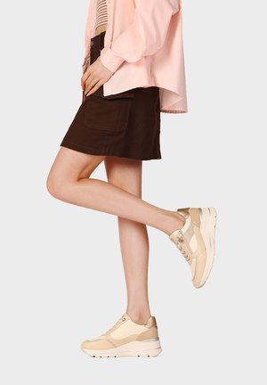 Persona che indossa sneakers beige, gonna marrone scuro con tasca, camicia rosa chiaro, posando con una gamba alzata su sfondo semplice.