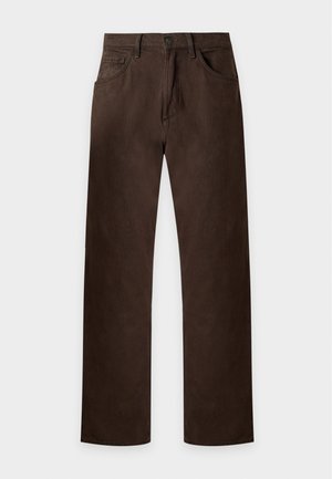 Jeune homme portant une chemise marron foncé à boutons, pantalon assorti, débardeur blanc et baskets blanches, debout avec une main dans la poche.