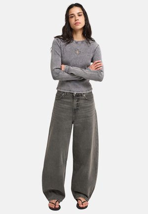 Jeune femme aux bras croisés portant un haut gris à manches longues côtelé avec une étoile et un jean noir délavé à jambes larges avec des sandales noires.