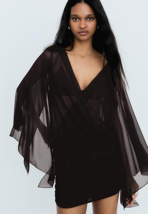 Vrouw met lang donker haar, gekleed in een zwarte doorschijnende jurk met brede vloeiende mouwen, staand tegen een effen lichte achtergrond.