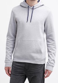 Mężczyzna w jasnoszarej bluzie z kapturem, z przednią kieszenią kangurką i ciemnymi sznurkami, w połączeniu z czarnymi dżinsami, na białym tle.