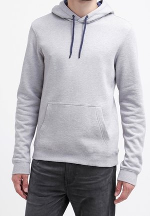 Mann trägt einen hellgrauen Kapuzenpullover mit einer vorderen Kängurutasche und dunklen Kordeln, kombiniert mit schwarzen Jeans, vor einem weißen Hintergrund.