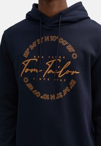 Sudadera azul marino de mezcla de algodón, con un gráfico redondo en dorado que muestra coordenadas y el nombre de la marca "Tom Tailor" destacado.