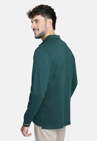 Camisa polo de manga larga verde con cuello, que presenta una textura suave y cortes laterales. Combinada con pantalones de color claro.