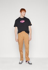 Črna majica s pink Nike logotipom, khaki cargo hlače in črne superge z rdečimi detajli. Model stoji pred belo ozadjem.