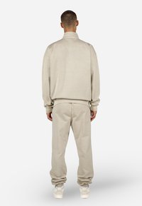 Bežs kokvilnas treniņtērps ar jaku ar rāvējslēdzēju un augstu apkakli, kā arī atbilstošām biksēm, abiem ir ribota apkakle un brīva piegriezuma siluets.