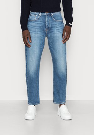 Man draagt lichtblauwe jeans met rechte pijpen, witte sneakers en een donker gekleurde trui met lange mouwen, staand tegen een effen achtergrond.