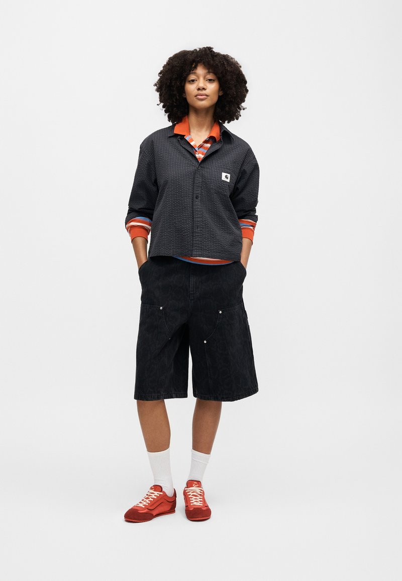 Junge Person mit lockigem Haar steht mit den Händen in den Taschen, trägt eine schwarze strukturierte Jacke, ein orange gestreiftes Hemd, schwarze lange Shorts, weiße Socken und rote Turnschuhe.