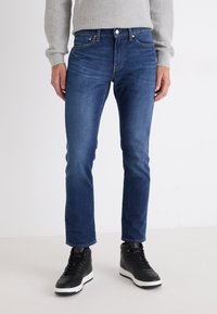 Mørkeblå jeans laget av denim, slim fit, med mid-rise midje, som har frontlommer, subtil søm, og en standard knapplukking.