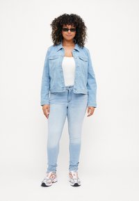 Chaqueta de mezclilla azul claro con dos bolsillos en el pecho, llevada sobre una blusa blanca. Combinada con jeans ajustados azul claro y zapatillas con un diseño estampado.