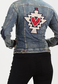 Widok z tyłu osoby w wyblakłej dżinsowej kurtce z czerwoną i białą łatką w kształcie serca i gwiazdy na plecach, zestawionej z czarnymi spodniami.