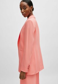 Peach blazer with a tailored design, notched lapels, open front, and welt pockets. Features button accents at the cuff for detail.