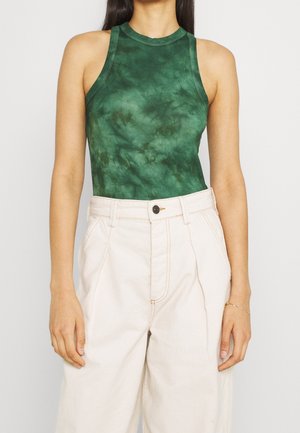 Woman wearing a fitted sleeveless dark green tie-dye top tucked into high-waisted off-white pants with a black button closure.
