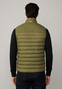 Olivgrüne Steppweste mit horizontaler Steppung, Reißverschluss vorne und hohem Kragen; getragen über einem schwarzen Langarmshirt und dunklen Jeans.