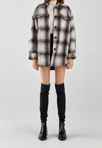 Kostkovaná bunda na knoflíky v černé a béžové barvě, měkký materiál, pohodlný střih, s dvěma kapsami na prsou, nošená přes krémový svetr a černé kraťasy.