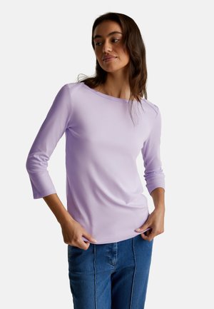 Vrouw in een lichtpaarse longsleeve top met 3/4 mouwen en blauwe jeans, staand met haar handen bij de taille en kijkend zijwaarts.