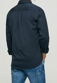 Maglietta blu navy a maniche lunghe con polsini arrotolati, dalla texture liscia e dettaglio di piega posteriore. Abbinata a jeans in denim azzurro chiaro.