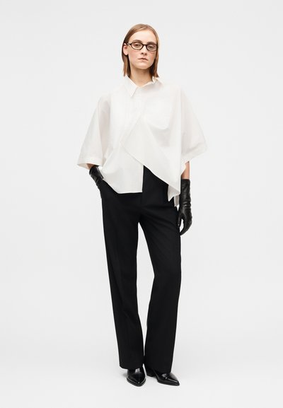 Άσπρο υπερμεγέθες πουκάμισο με τσέπη στο μπροστινό μέρος, συνδυασμένο με ίσια μαύρη παντελόνια. Το μοντέλο φοράει μαύρα δερμάτινα γάντια και ψηλοτάκουνα παπούτσια με μυτερή μύτη.