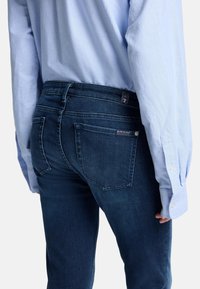 Mörkblå denimjeans med en figurnära design, fem fickor och en logotypetikett i bakänden av midjan. Kombineras med en ljusblå långärmad skjorta.