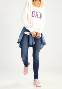 Weißes Sweatshirt mit lila "GAP"-Buchstaben, blaue Jeansjacke um die Taille gebunden, blaue enge Jeans und rosa Sneakers.