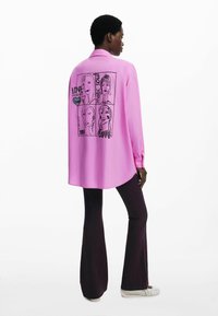 Roze langemouwen shirt met een gebogen zoom, voorzien van een zwart grafische illustratie op de achterkant met gezichten en tekst. Gecombineerd met donkere flared broeken.