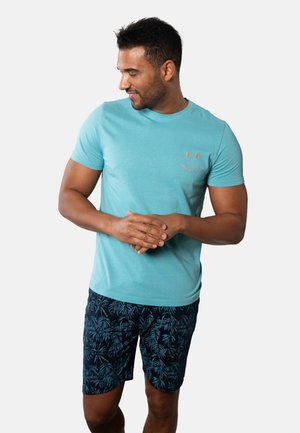 Man wearing a turquoise t-shirt with a small orange smiley face and dark blue shorts with a leaf pattern, looking down and to the side.