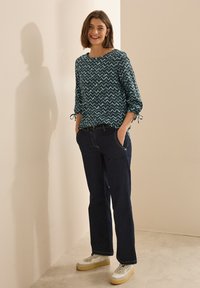 Langärmlige, tealfarbene Bluse mit Zickzackmuster, kombiniert mit dunklen Denim-Jeans und weißen Sneakers mit dicken Sohlen.