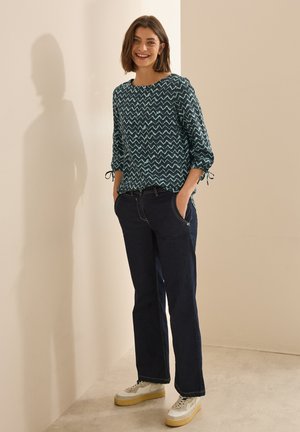 Blouse à manches longues de couleur turquoise avec un motif à zigzag, associée à un jean en denim noir et des baskets blanches à semelles épaisses.