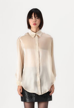 Satin blouse in light beige with a collared neck, long sleeves, front buttons, and a flowing silhouette; worn with a patterned black skirt.