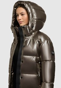 Puffet dunjakke i glat brunt stof. Har en stor hætte, trykknaplukning, sidelommer og et logo-patch på ærmet.