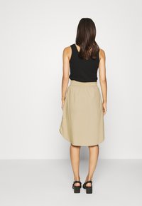 Rok midi beige dengan waistband elastis, kain mengalir, dan hem sedikit melengkung. Dipasangkan dengan atasan tanpa lengan hitam dan sandal bertumit block.