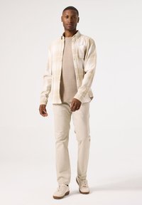 Licht geruit overhemd met een knoopsluiting, beige trui eronder, licht crèmekleurige broek en beige sneakers met bruine accenten.