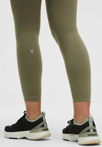 Olíva zöld leggingset és fekete-fehér Lululemon tornacipőt viselő lábak, hátulról, fehér háttér előtt.