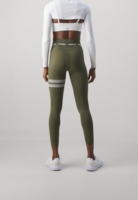 Leggings vert olive à la texture lisse, ornés de rayures horizontales blanches sur la cuisse et d'une ceinture avec logo. Complétés par des baskets blanches.