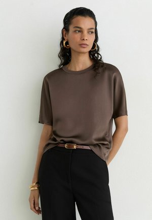 Femme portant un haut marron à manches courtes, un pantalon noir avec une ceinture marron, des boucles d'oreilles en anneaux dorés et des bracelets dorés, debout contre un mur blanc.