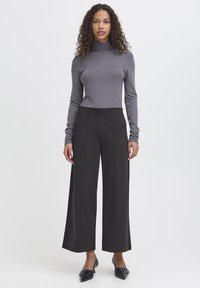 Kobieta o kręconych włosach, ubrana w szary golf, czarne szerokie spodnie do połowy łydki i czarne buty na szpic, stojąca na białym tle.