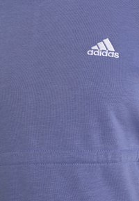 Camiseta morada de tela suave, con un logo de Adidas bordado en blanco en la parte superior izquierda. Tiene una textura lisa y un diseño sencillo.