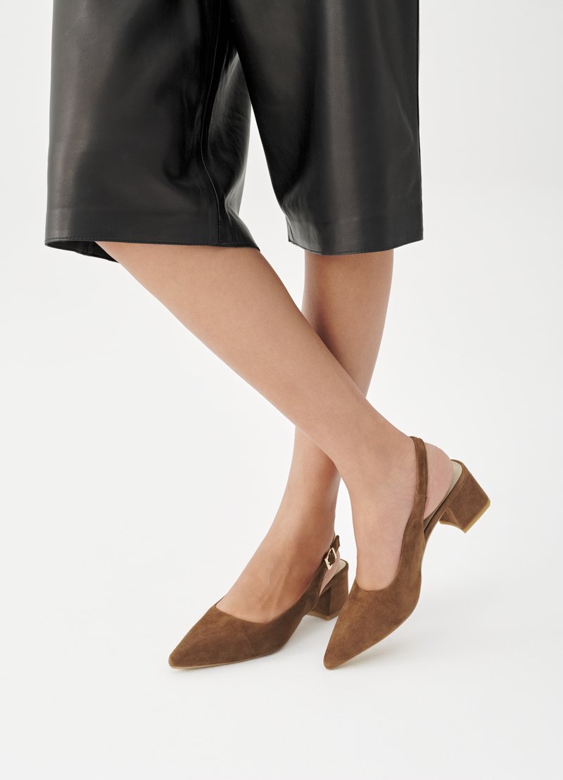 Beine übereinandergeschlagen, getragen braune Wildleder-Slingback-Schuhe mit Blockabsatz und schwarze kniehohe Lederhosen vor weißem Hintergrund.