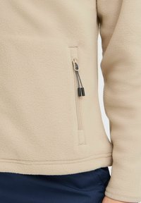 Beige fleece takki, jossa on teksturoitu pinta, sivussa vetoketjulla varustettu tasku ja mustat vetoketjun vetimet, suunniteltu lämpöä ja mukavuutta varten.