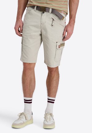Man die beige cargoshorts met riem draagt, een beige-wit gestreept hemd, witte sokken met strepen en witte sneakers, staand tegen een witte achtergrond.