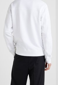 Personne portant un sweat-shirt blanc uni à manches longues et un pantalon noir, vue de dos sur un fond clair uni.