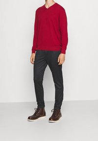 Un pull en maille rouge à col en V associé à un pantalon gris foncé, ajusté, et des bottes en cuir marron à lacets avec une semelle blanche, le tout sur un fond uni.