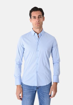 Giovane uomo con capelli scuri che indossa una camicia azzurro chiaro abbottonata e jeans blu, in piedi davanti a uno sfondo chiaro uniforme.