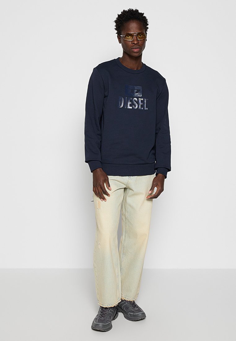 Felpa blu navy con logo "DIESEL" in rilievo, maniche lunghe, polsini a costine, abbinata a jeans chiari manomessi e sneakers grigie.