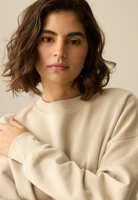 Svetlo bež pulover od mekanog pamučnog materijala. Ima okrugli izrez i opušten kroj, s rebrastim manšetama i suptilnim detaljima šava.