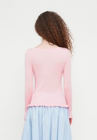 Γυναίκα με καστανά σγουρά μαλλιά, που φορεί ένα ριμπ ροζ ανοιχτό ροζ μακρυμάνικο τοπ και μια ανοιχτομπλε  πλισέ φούστα, όρθια με την πλάτη γυρισμένη.