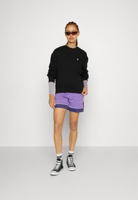 Schwarzer Sweatshirt mit grauen Bündchen, lila Shorts mit weißen Paspeln, schwarze High-Top-Sneaker und reflektierende Sonnenbrille.