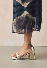 Arany színű, kígyóbőr mintázatú blokk sarokkal ellátott szandál bokapánttal. Nyitott orrú kialakítás, széles elejű pántokkal. Különféle alkalmakra alkalmas.