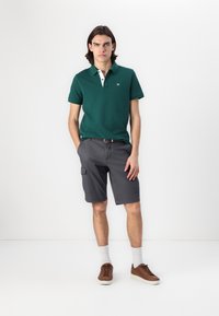Zöld póló fehér gallérral, szürke cargo sorttal párosítva. Barna sneakers és fehér zoknik egészítik ki az öltözéket. Semleges háttér.